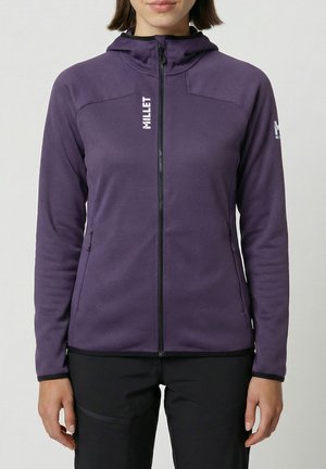 Femme portant un sweat à capuche zippé violet avec le logo "MILLET", pantalon noir, debout devant un fond clair uni.