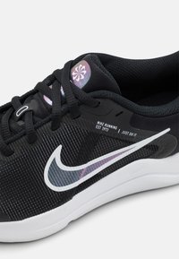 Chaussure de sport noire avec une tige en mesh texturé, logo swoosh Nike blanc et un accent réfléchissant sur le col. Équipée de lacets noirs.
