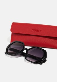 Ochelari de soare hexagonali negri cu lentile gradient alături de un toc din piele roșie embosat cu "GUESS."