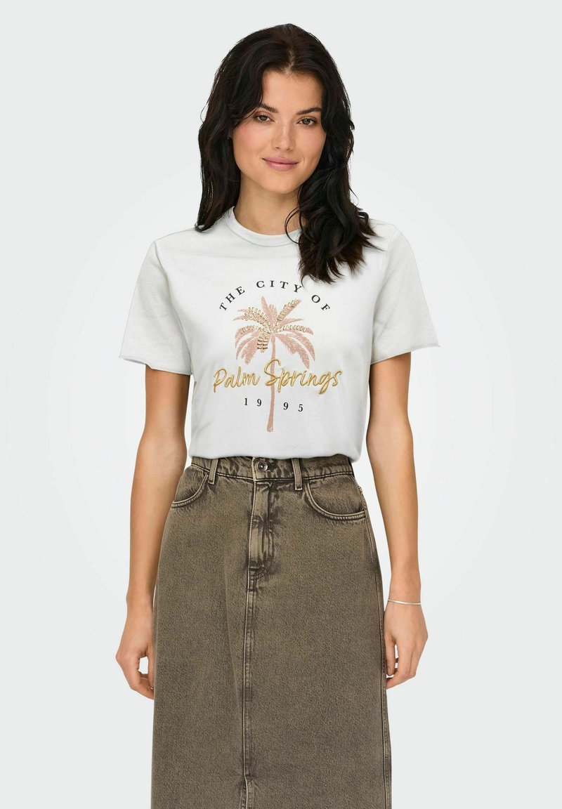Camiseta blanca de algodón con un gráfico que dice "La Ciudad de Palm Springs" que presenta palmeras en rosa y dorado, combinada con una falda de mezclilla marrón de cintura alta.