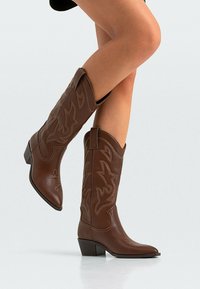 Ben som bär bruna lädercowboyboots i midjehöjd med dekorativ sömnad, ett ben upphöjt, mot en ljus, neutral bakgrund.