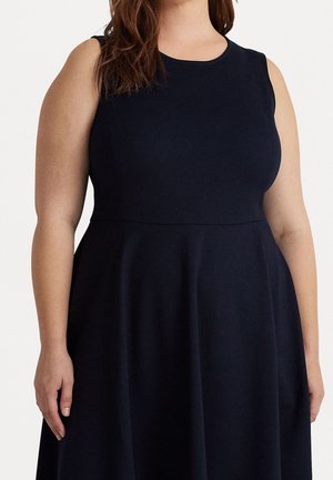 Vrouw in een mouwloze zwarte jurk met nauwsluitend lijfje en uitlopende rok, bruin haar rustend op de schouder, lichte huidtint, stilstaand.