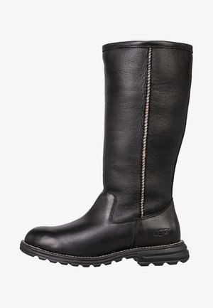 Bota negra de cuero hasta la mitad de la pantorrilla con suela resistente, costuras laterales visibles y logo UGG en relieve en el panel del talón.