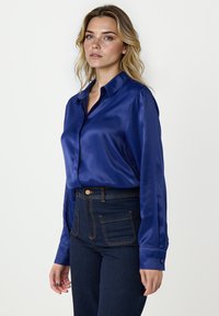 Niebieska bluzka satynowa z wysmuklonym kołnierzem, z przodu zapinana na guziki, z długimi rękawami. Zestawiona z ciemnoniebieskimi dżinsami z wysokim stanem, ozdobionymi złotymi szwami.