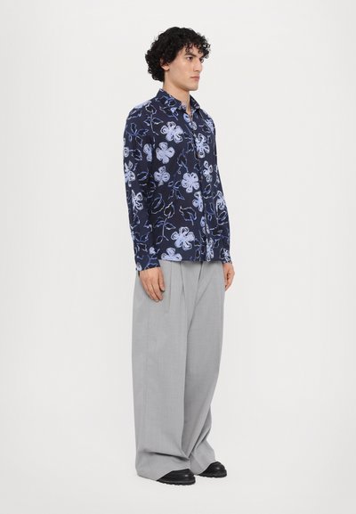 Camisa de botones floral en color navy con patrones en blanco y azul claro, de mangas largas, combinada con pantalones grises de pliegues y zapatos negros.