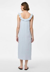 Gaiši zila bez piedurknēm kleita ar volāniem pie kakla un teksturētu audumu. Ankle garuma, ar brīvu piegriezumu, valkāta ar melniem čībām.