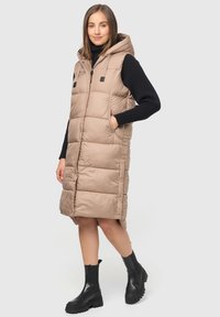 Gilet matelassé beige avec capuche, présentant une fermeture éclair sur le devant, des poches latérales et des accents noirs. Porté par-dessus un pull côtelé noir et des bottes noires chunky.