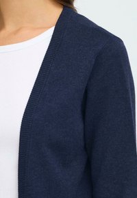 Cardigan marine fabriqué en tissu tricoté. Design à front ouvert avec des bords côtelés. Superposé sur un haut à col rond côtelé blanc.