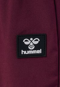 Bordeaux-textil mit einer weichen Textur, die ein schwarzes rechteckiges Etikett zeigt, das ein weißes Logo und den markant eingeprägten Markennamen "hummel" trägt.