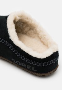 Svart mockasinskon med mjuk tanfleecefoder, sydd detaljer längs öppningen och "SOREL"-logotyp tryckt på sidan.