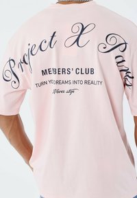 Persona luciendo una camisa rosa pastel con texto negro: "Project X Paris", "Club de miembros", "Convierte tus sueños en realidad" y "Nunca te detengas".