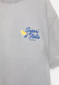 Szürke pamut póló kerek nyakkal. Kék színű "Sapori d'Italia" felirattal és egy sárga citrom grafikával rendelkezik. Hétköznapi, laza szabású.