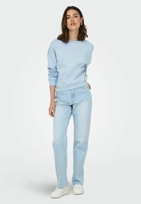 Lichtblauwe crewneck sweatshirt met afgeknipte mouwen, gecombineerd met lichtgekleurde rechte jeans en witte sneakers. Eenvoudige, casual look.