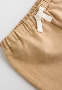 Pantalon à taille élastique beige avec un cordon en tissu blanc noué en nœud sur un fond blanc.