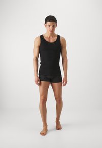 Čierny bezrukávový top a tmavé boxerky na mužskom modeli, ktorý stojí naboso pred jednoduchým bielym pozadím. Hladká, priliehavá tkanina.