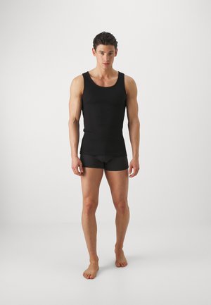 Schwarzes ärmelloses Tanktop und dunkle Boxershorts an einem männlichen Modell, das barfuß vor einem schlichten weißen Hintergrund steht. Glatter, enganliegender Stoff.