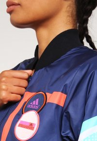 Navy bomber jacket with black collar, pink and aqua stripes, featuring circular Adidas patches in pink and black on the chest.