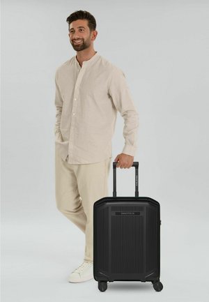 Hombre con atuendo beige sonriendo, de pie mientras sostiene una maleta negra con ruedas SMARTBOX por el asa.