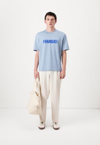 Light blue cotton T-shirt with "HUGO" in darker blue text, paired with cream wide-leg trousers and a beige tote bag. Casual footwear completes the look.