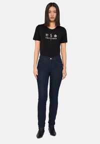 Camiseta de algodón negra con logo impreso, combinada con unos pantalones vaqueros de talle alto de color azul oscuro. El modelo lleva botas de tobillo negras. Fondo liso.