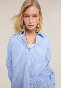 Joven con cabello rubio que lleva una camisa azul a rayas con bordado floral y joyas de plata sobre un fondo liso.