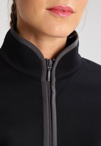Giacca nera con zip e colletto alto, caratterizzata da una cerniera e bordi grigi. Tessuto morbido e design aderente in evidenza.