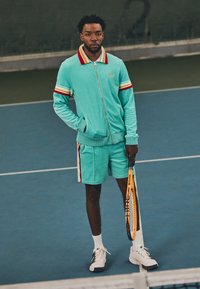 Veste de sport vert menthe et shorts avec des accents rayés en rouge et jaune. Chaussures de tennis blanches, tenant une raquette de tennis. Cadre intérieur de court.