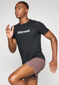 T-shirt de sport noir en tissu texturé avec le logo "Normal" en blanc ; le modèle est en mouvement portant un short marron avec des fentes sur les côtés.