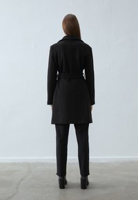 Svart frakk med belte i midjen, lange ermer og rett silhuett. Materialet ser glatt ut med en matt finish.