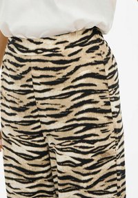 Pantalons rayés tigre beiges et noirs avec une taille haute, présentant une texture lisse et une coupe large. Pas de matériel ou d'accents visibles.