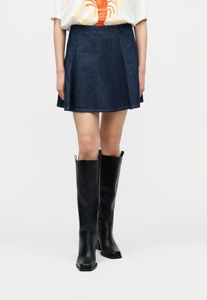 Femme portant une mini-jupe en denim foncé, un T-shirt blanc avec une impression de homard orange, et des bottes en cuir noires montantes jusqu'aux genoux, debout devant un fond blanc.