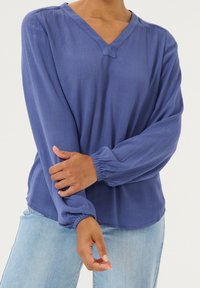 Blouse bleue à manches longues avec un col en V et des poignets froncés ; tissu léger à texture subtile ; portée avec un jean denim clair.