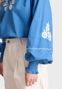 Blusa di cotone blu con maniche lunghe e puffate, caratterizzata da ricami floreali bianchi ai polsi. Abbinata a pantaloni chiari a pieghe.