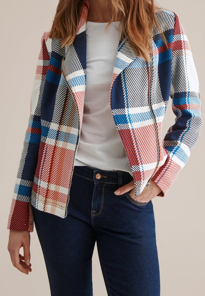 Femme portant une veste zippée à carreaux colorés sur une chemise blanche et un jean bleu foncé, avec une main dans la poche.