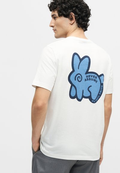 Νέος άνδρας με σγουρά μαλλιά που φοράει λευκό μπλουζάκι με μεγάλο μπλε καρτούν λαγό και τη φράση «Never Assume» στην πλάτη.