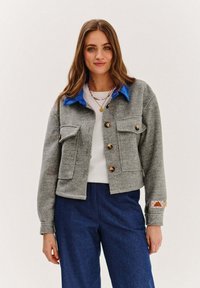 Sivý vlnený kabát so zmesou s modrým zamatovým golierom, gombíkovým zapínaním a dvoma náprsními vreckami. Obsahuje prešívaný vzor na akcentoch manžiet.