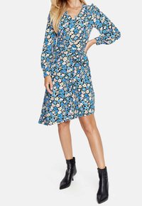 Modelka w kwiecistej sukience z długimi rękawami w odcieniach niebieskiego, różowego i żółtego, zestawiona z czarnymi botkami za kostkę, stojąca pewnie.