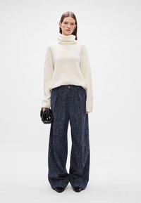 Pulôver de gola alta creme com um tricot texturizado, combinado com calças largas de tweed azul-marinho. Bolsa preta com um pequeno detalhe de fecho.