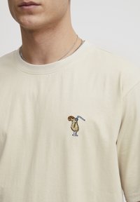 Beige cotton t-shirt featuring a small embroidered design of a drink in a glass, with a straw and a slice of fruit on top.