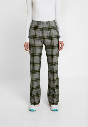 Personne portant un pantalon à carreaux gris avec des rayures vertes et jaunes, un haut blanc à manches longues et des baskets blanches à semelles bleues et roses.