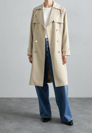 Personne portant un trench-coat beige croisé, un haut blanc, un jean bleu à jambes larges et des chaussures noires, debout sur un sol gris contre un mur clair.