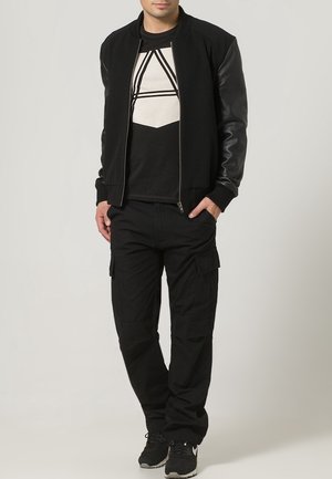 Black bomber jacket with leather sleeves, layered over a black graphic t-shirt featuring geometric white design, paired with black cargo pants.