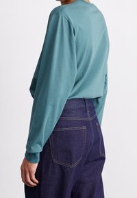 Tealfärgad långärmad t-shirt med rund halsringning, kombinerad med högmidjade mörkblå jeans med gula sömmar och bakfickor.