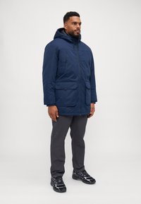 Chaqueta con capucha de color azul marino, con cierre de cremallera, dos bolsillos frontales y cordones ajustables. Combinada con pantalones grises y zapatillas negras.