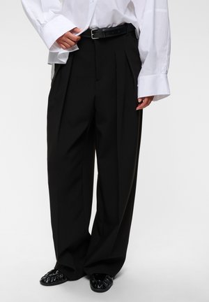 Personne portant un pantalon plissé noir à jambes larges, une chemise blanche oversize, une ceinture noire et des chaussures plates noires brillantes, debout devant un fond uni.
