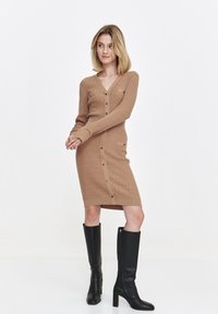 Sukienka dziana w kolorze beżowym z dekoltem w kształcie V, długimi rękawami, zapinana na guziki z boku i z kieszeniami. Połączona z wysokimi czarnymi botkami na obcasie. Gładka faktura.
