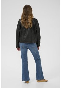 Melnā ādas jaka ar brīvu piegriezumu un rāvējslēdzēja aprocēm, kombinēta ar gaišziliem platām biksēm un bēšām kurpēm. Skats no muguras.