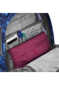 Purple fabric pencil case with a textured design, located inside a gray backpack. Blue floral pattern on the backpack's exterior visible.