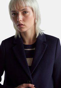 Navy kabát vyrobený z vlněné směsi s klasickým límcem, zapínáním na jeden knoflík a měkkou texturou. Naležený přes pruhovaný pletený svetr.