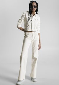 Chaqueta de denim blanca y jeans acampanados, con un diseño cropped, botonera frontal y tela texturizada. Combinados con zapatos grises y gafas de sol.